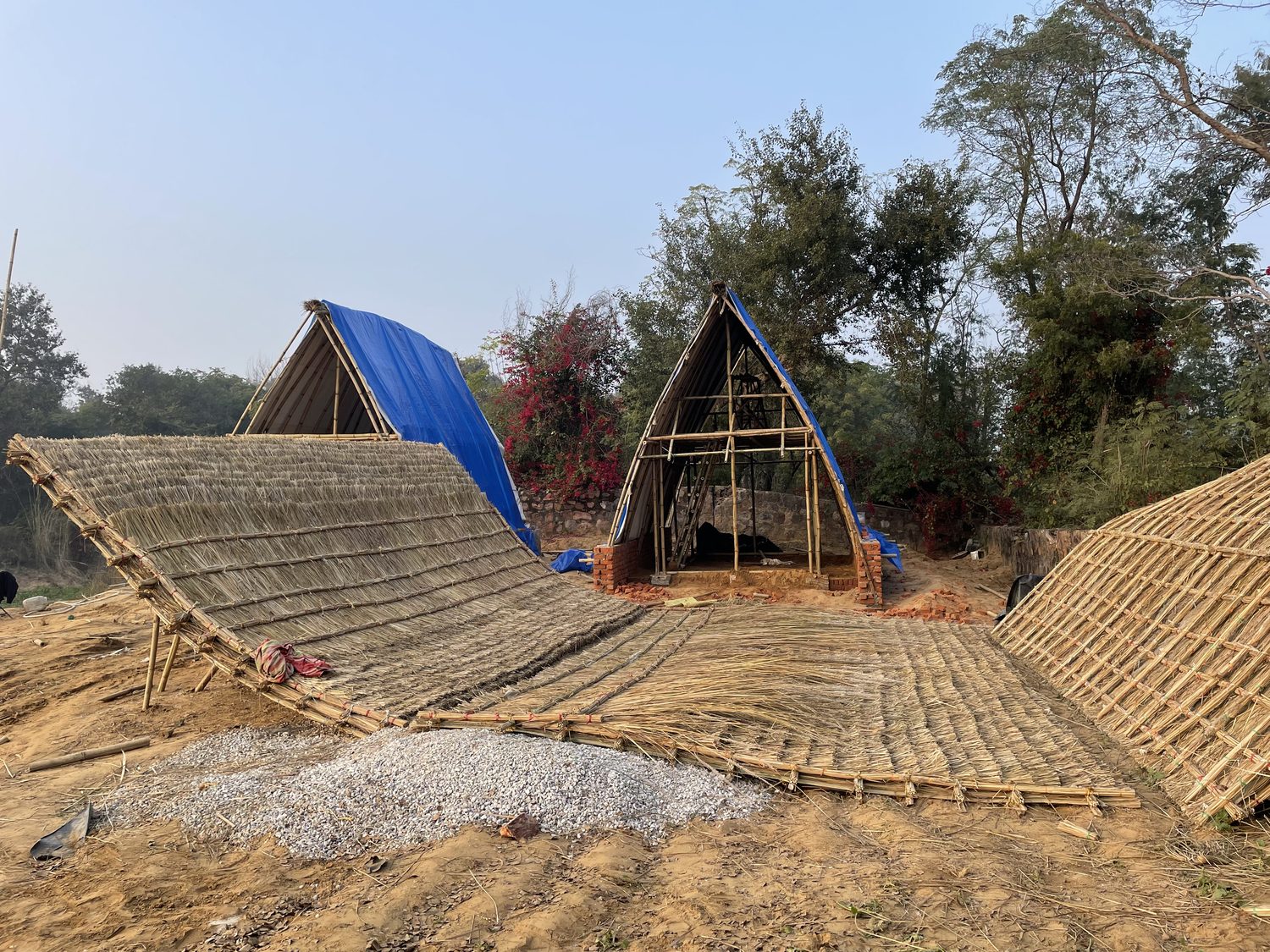 Large woven thatch panels laid flat on gravel and earth in front of two bamboo frames