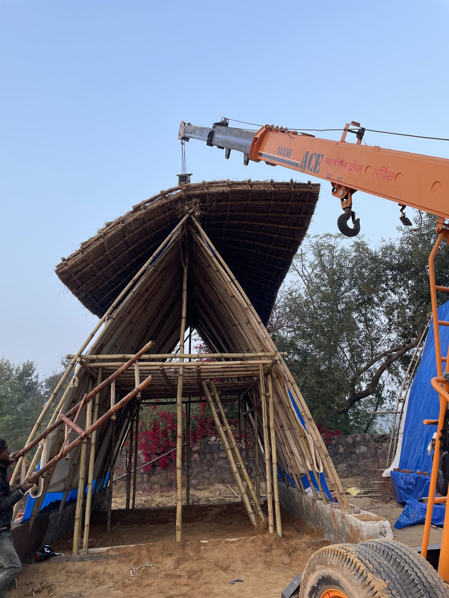 A mobile crane lifting a curved thatch panel through the air onto a bamboo A-frame