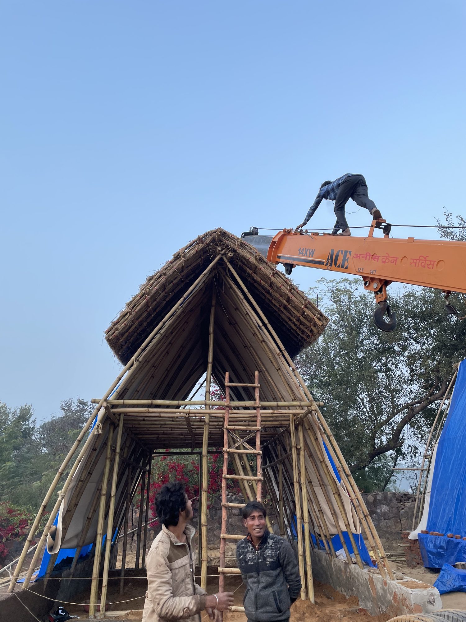 A man standing on the ridge of a thatched A-frame against a clear sky, finishing the apex