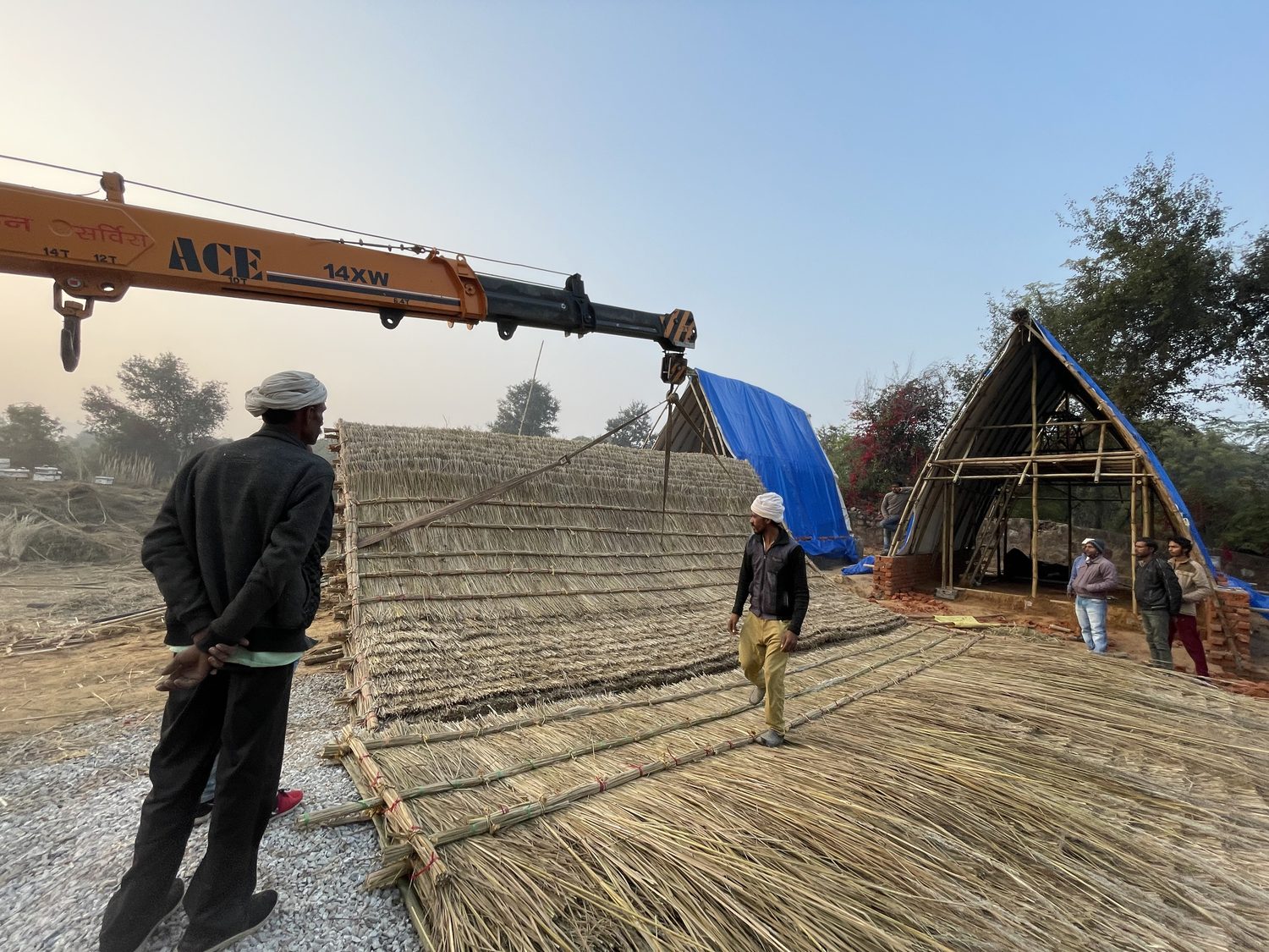 Two men guiding a large thatch panel on the ground while a crane boom reaches over them