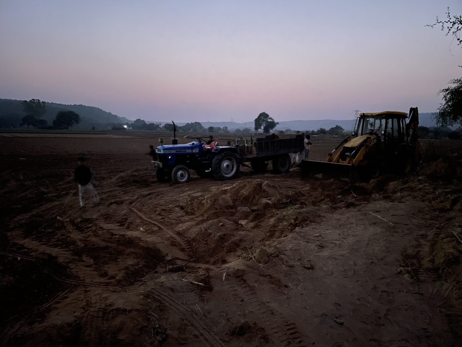 A tractor and a JCB standing on freshly turned earth at dusk, a man beside them, hills on the horizon
