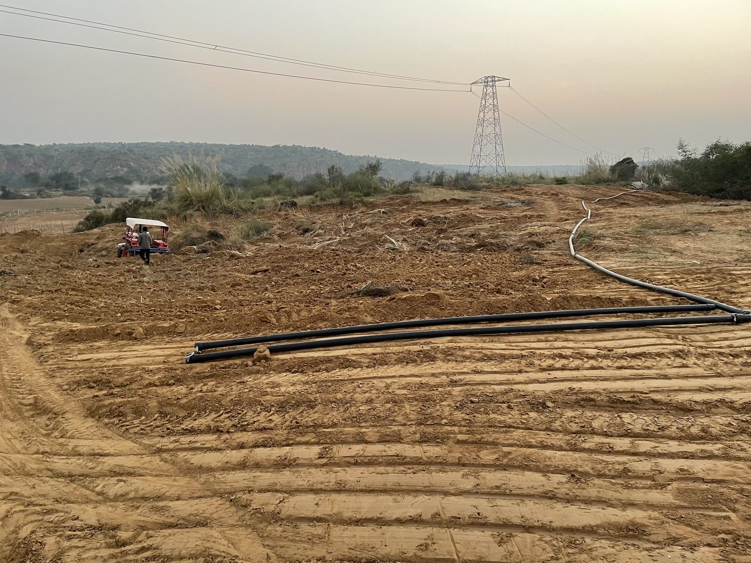 Lengths of black irrigation pipe laid out across bare worked earth, a transmission pylon and hills beyond