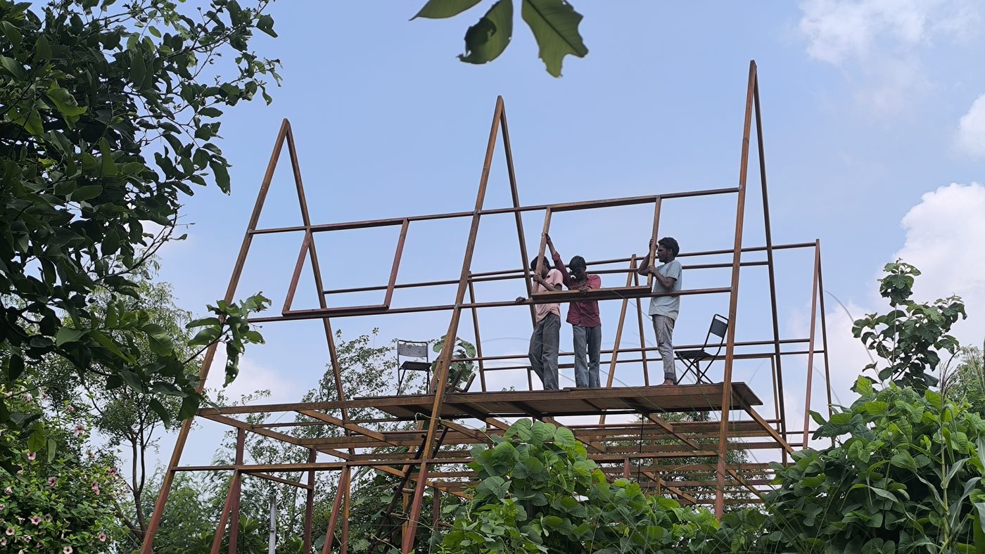 Two men standing on a steel A-frame under construction, green trees behind, monsoon sky