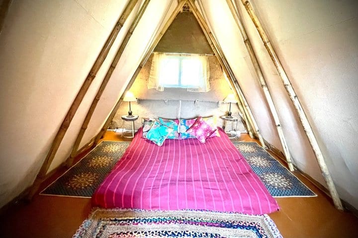 Inside an A-frame mud house: a bamboo frame rising to a point, a low bed, two lamps and a small window