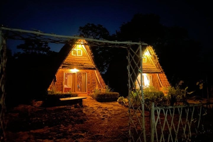 The two A-frame houses lit from inside at night, seen through a bamboo gate