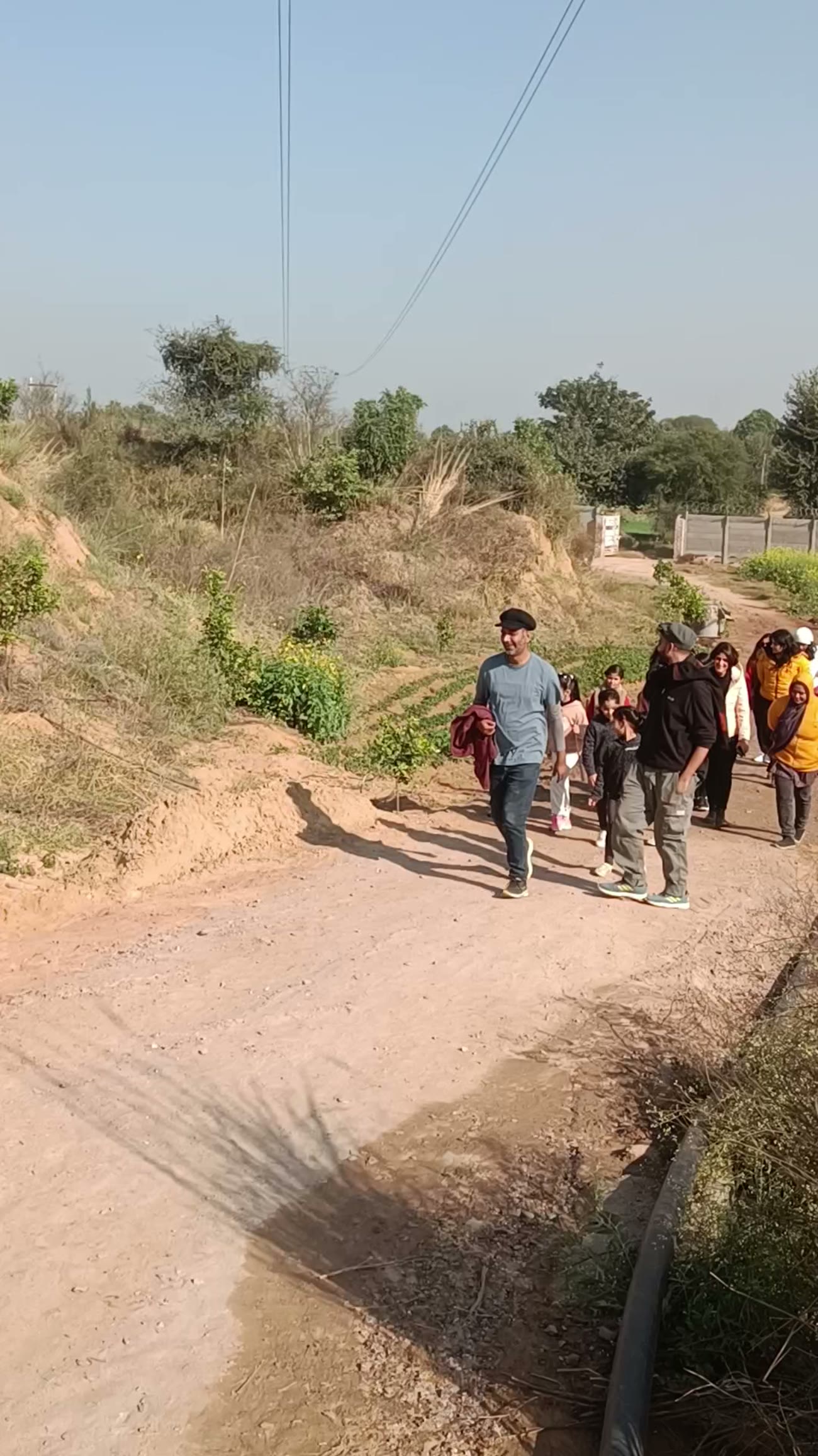 A line of children and adults walking up a sandy track towards the farm, scrub and a boundary wall either side