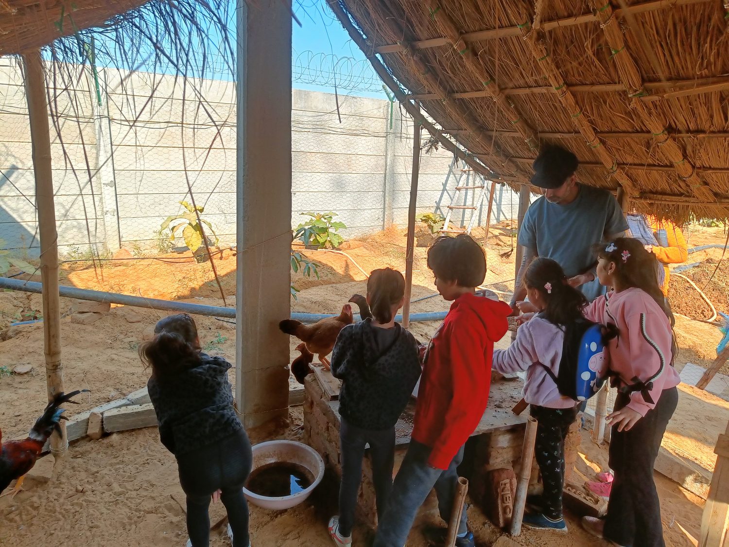 Children gathered along a trough under a thatched shelter, hands reaching in, an adult working alongside them