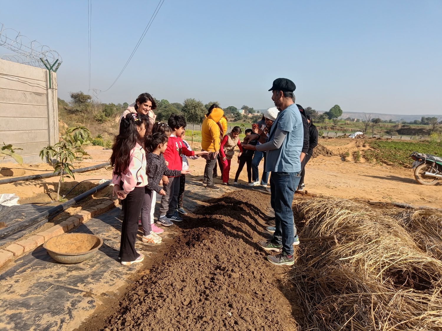 Children and two adults standing around a long compost heap of dark material with straw beside it