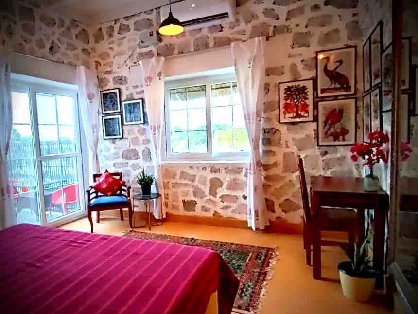 A bedroom with a pink bedspread, stone walls hung with framed prints, and glass doors onto a balcony