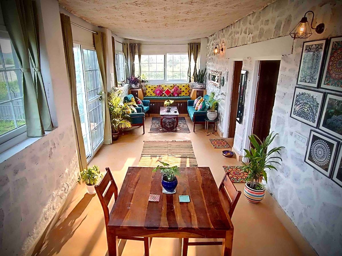 A wooden dining table in a stone room, framed folk-art prints on the walls, light from windows on both sides