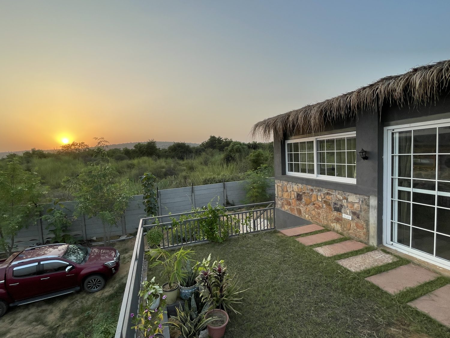 A stone house under a thatched roof at sunrise, glass doors opening onto a lawn, stepping stones across the grass