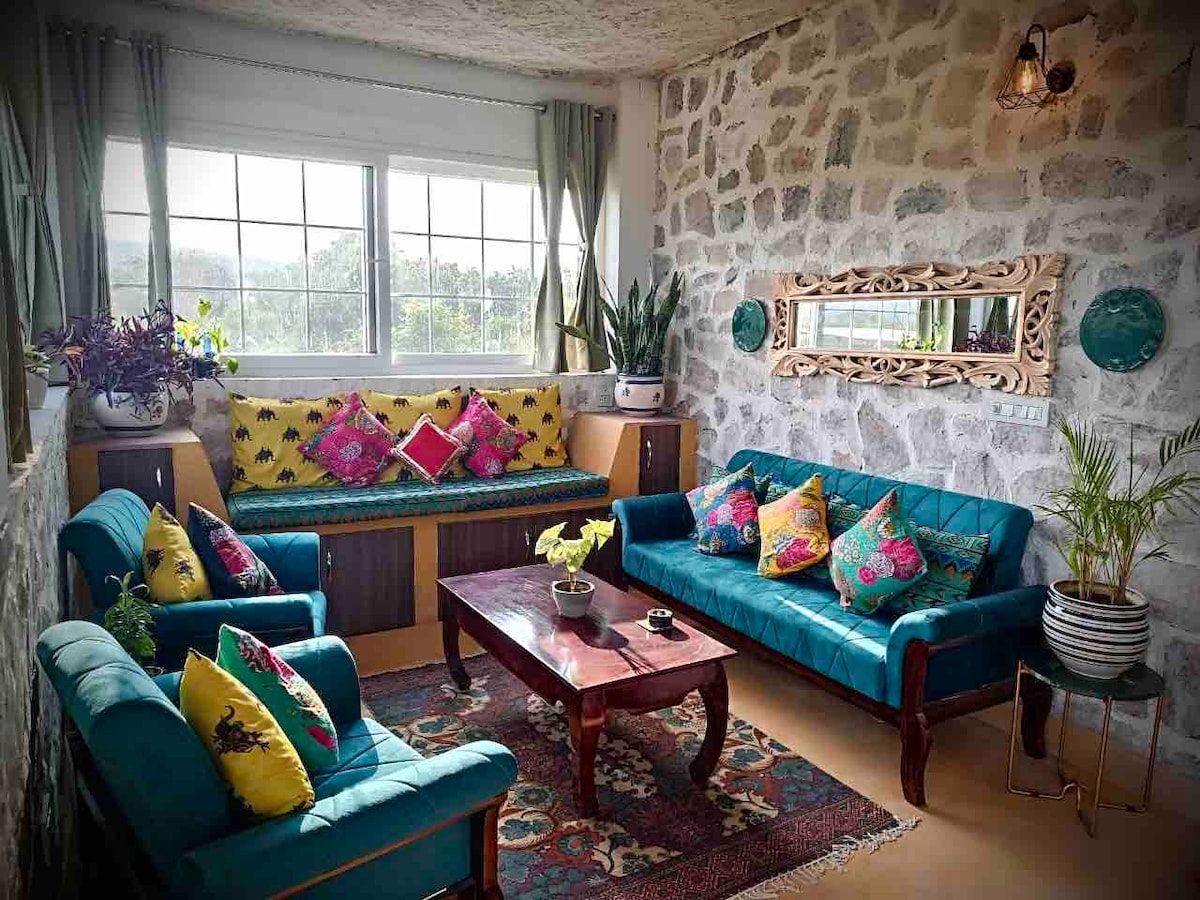 A sitting room with teal sofas and bright block-printed cushions against a rough stone wall, a mirror and plants